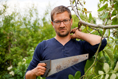 Portrait of man holding hand saw while standing against tree