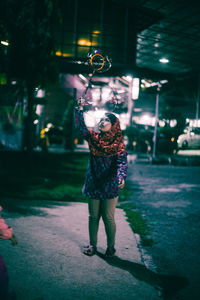 Full length of girl playing with bubbles while standing on footpath at night