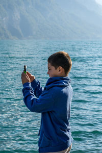Side view of boy photographing through mobile phone against sea