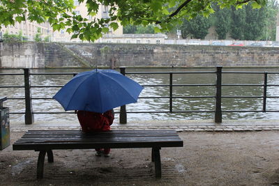 Rear view of boy with umbrella sitting on bench against canal