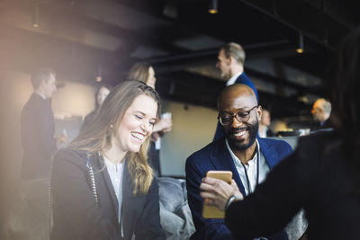 Smiling male and female coworkers looking at mobile phone while sitting with entrepreneur in office