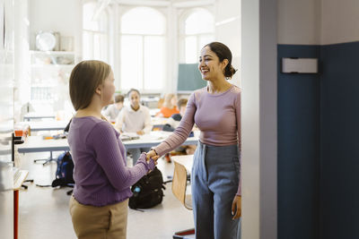 Smiling female teacher doing handshake with student standing in classroom