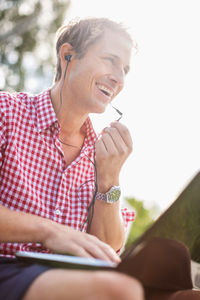 Low angle view of happy man using hands-free device against clear sky