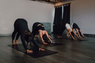 Fit barefoot unrecognizable women in sportswear concentrating and doing downward facing dog exercise on sports mats on wooden floor against white walls of spacious hall