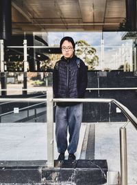 Portrait of young man standing at railings against reflective building facade.