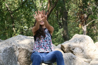 Happy woman showing stop gesture while sitting on rock against trees