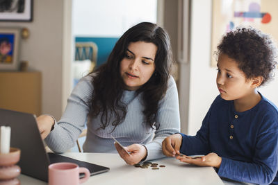 Son counting coins while sitting with mother using laptop at home