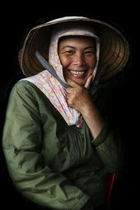 Portrait of smiling mature woman wearing asian style conical hat against black background