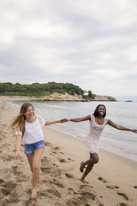 Cheerful female friends holding hands while running on beach against sky