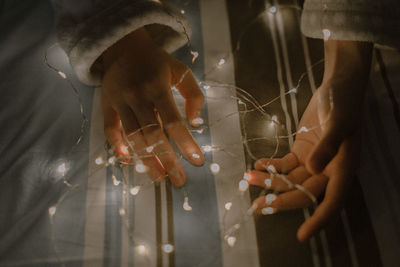 High angle view of hands with illuminated string lights on bed