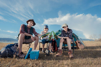 Portrait of friends toasting beer bottles while camping on grassy field against blue sky