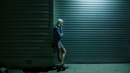 Full length of sad young woman standing by closed shutter at night