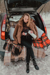 Blonde woman wrapped in blanket in trunk car drinking coffee tea from thermos. travel in winter. car 