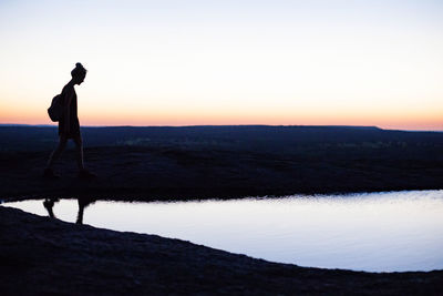 Side view of silhouette woman walking by pond on field during sunset