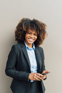 Happy businesswoman holding smart phone standing in front of wall