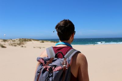 Rear view of young man with backpack standing at beach against blue sky during sunny day