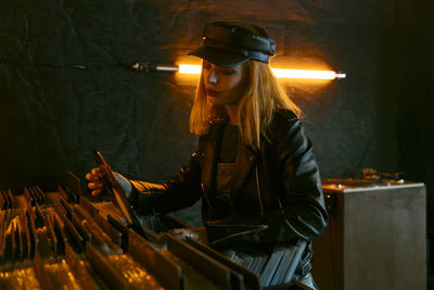 Serene young female in black cap choosing vinyl record while standing near counter with collection of musical albums in store with glowing lamps