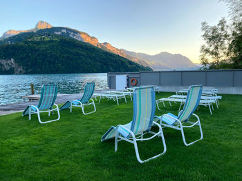 Empty chairs on grass by lake against sky