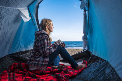 Woman sitting in tent at beach against sky