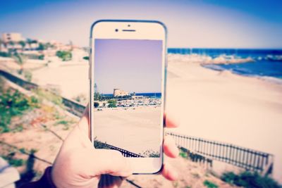 Close-up of hand holding mobile phone at beach against sky