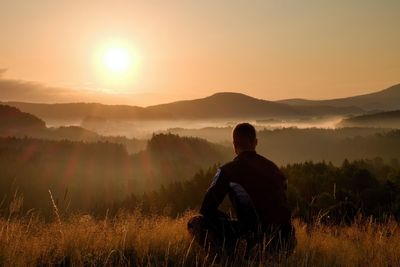 Hiker in squatting position in high grass meadow enjoy the colorful sunrise scenery