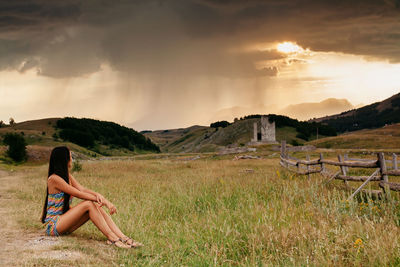 Side view of woman looking at mountains while sitting on field