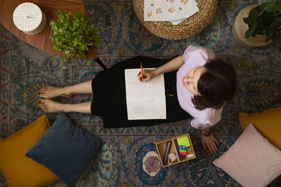Full body adult female artist sitting on floor near table with laptop and drawing sketch in sketchbook at home