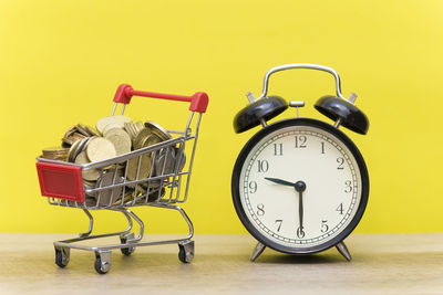 Close-up of alarm clock by coins in shopping cart on table against wall
