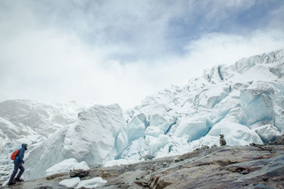 Side view of hiker walking on field by snow covered mountain against cloudy sky