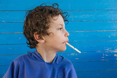 Close-up of boy eating popsicle looking away against blue wall