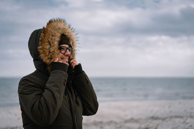 Man with hooded jacket standing at beach against sky during winter