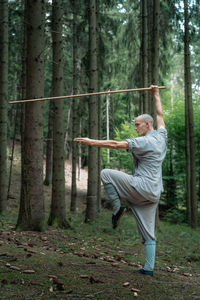 Full body bald man in traditional garments practicing with bamboo stick during kung fu training in coniferous woods