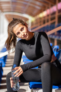 Fit young woman in black sportswear resting after workout or running, looking at camera.