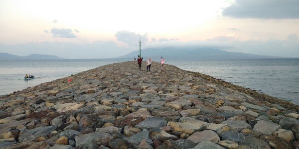 People on rocks at beach against sky during sunset