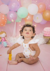 Cute baby girl with balloons and birthday cake sitting at home