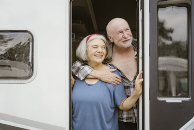 Smiling senior man hugging woman from behind while standing at motor home doorway