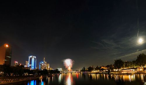 Illuminated cityscape in front of river against sky at night