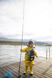 Baby boy wearing raincoat and life jacket while holding fishing rod on wooden pier over lake