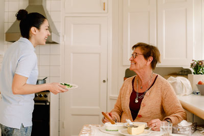 Happy elderly woman talking with female healthcare worker in kitchen at nursing home