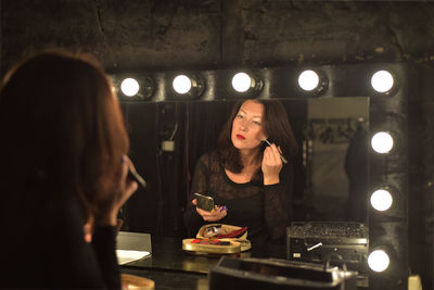 Young woman applying make-up reflecting on mirror in illuminated room