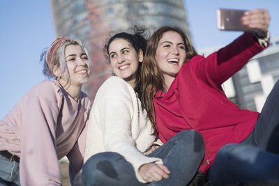 Happy female friends taking selfie while sitting on building terrace against sky