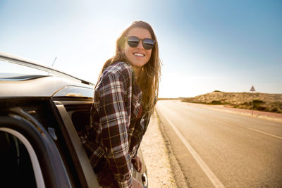 Portrait of young woman wearing sunglasses while peeking out from car on road against sky