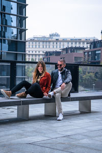 Happy friends sitting on seat against buildings in city