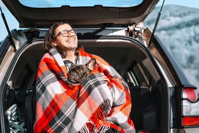 Portrait of smiling mature man and french bulldog sitting in car trunk in winter landscape