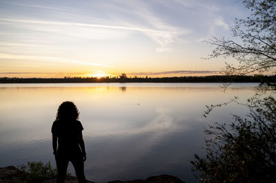 Rear view of woman standing by lake against sky during sunset