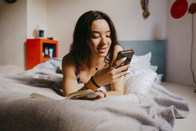 Smiling young woman using smart phone while lying on bed at home