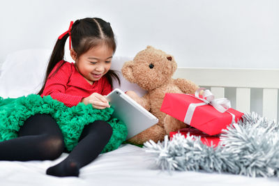 Cute girl using digital tablet while sitting with christmas gift and teddy bear in bed at home