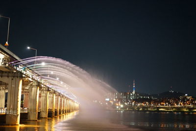 Illuminated modern buildings by river against sky at night