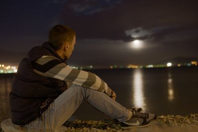 Side view of young man sitting at beach against sky at night