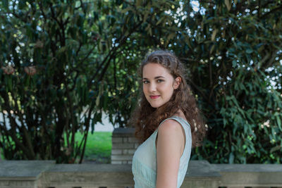 Beautiful curly hair young woman in light blue simple dress on balcony, park trees background